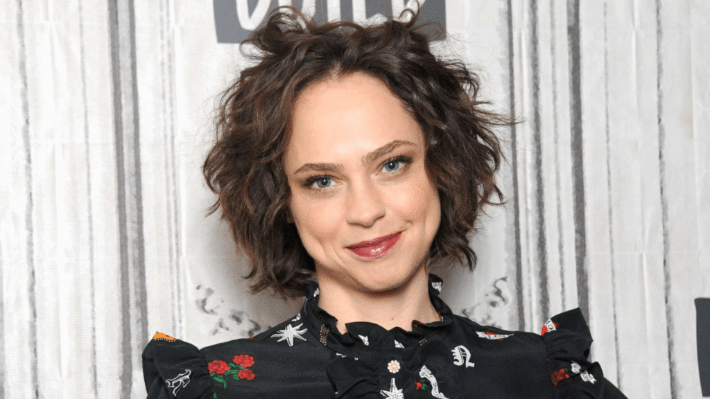 kristina in a patterned black blouse, smiling with short curly hair, standing against a textured backdrop